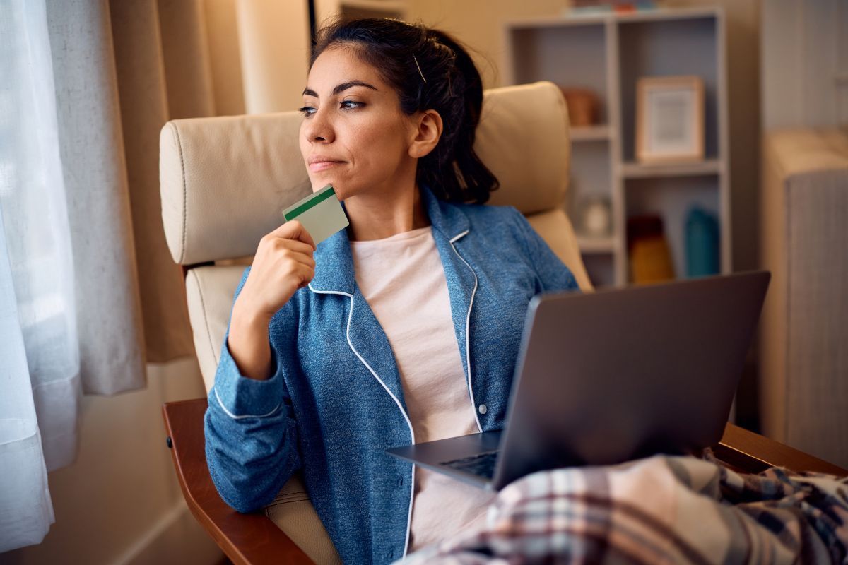 woman on a laptop looking out the window with a credit card in her hand thinking