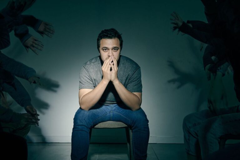 Man looking worried sitting in a chair, background shadows of hands all around him reaching for him