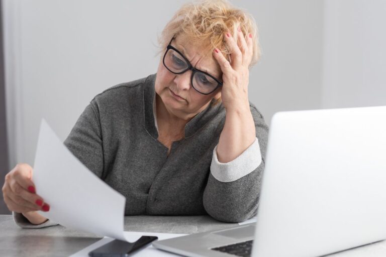 Older woman looking at a paper looking confused infront of her laptop