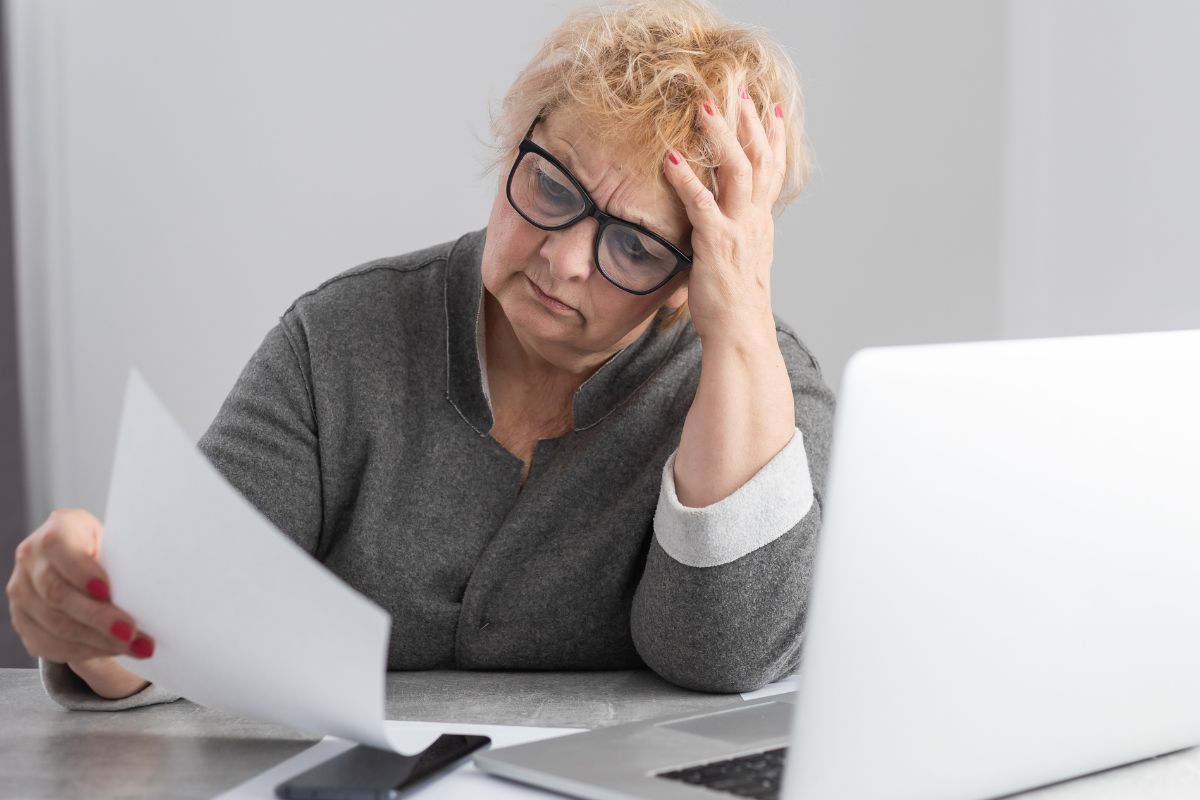 Older woman looking at a paper looking confused infront of her laptop