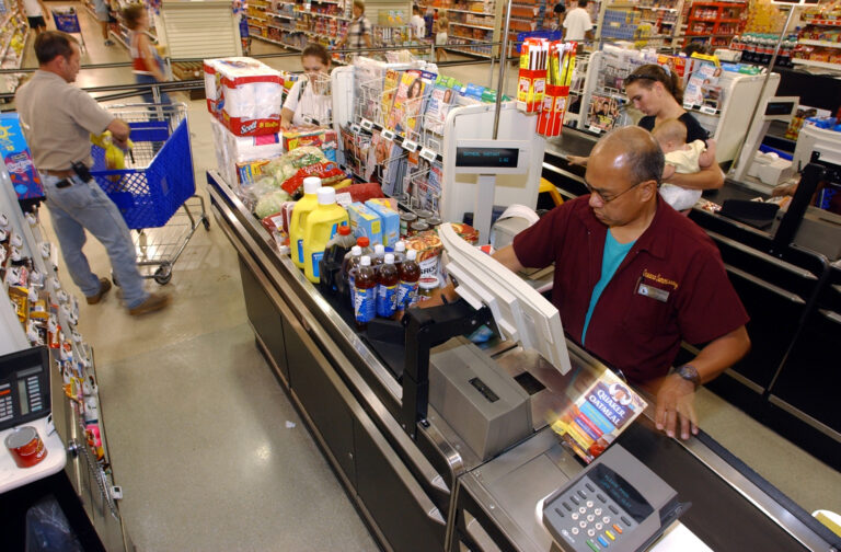 A family unloads groceries from a shopping cart at a store checkout counter