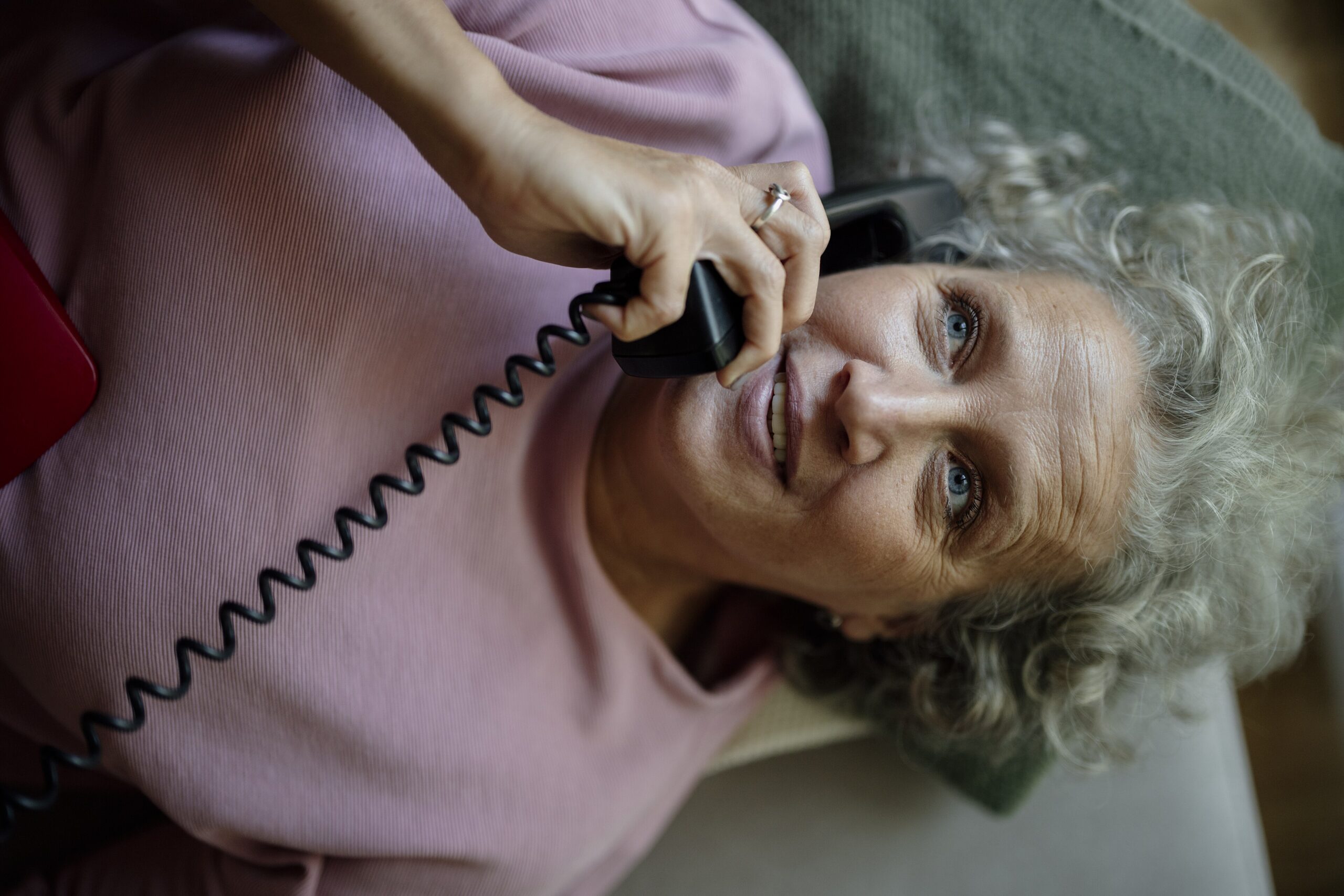 An older woman talking on a landline phone while resting on a couch at home